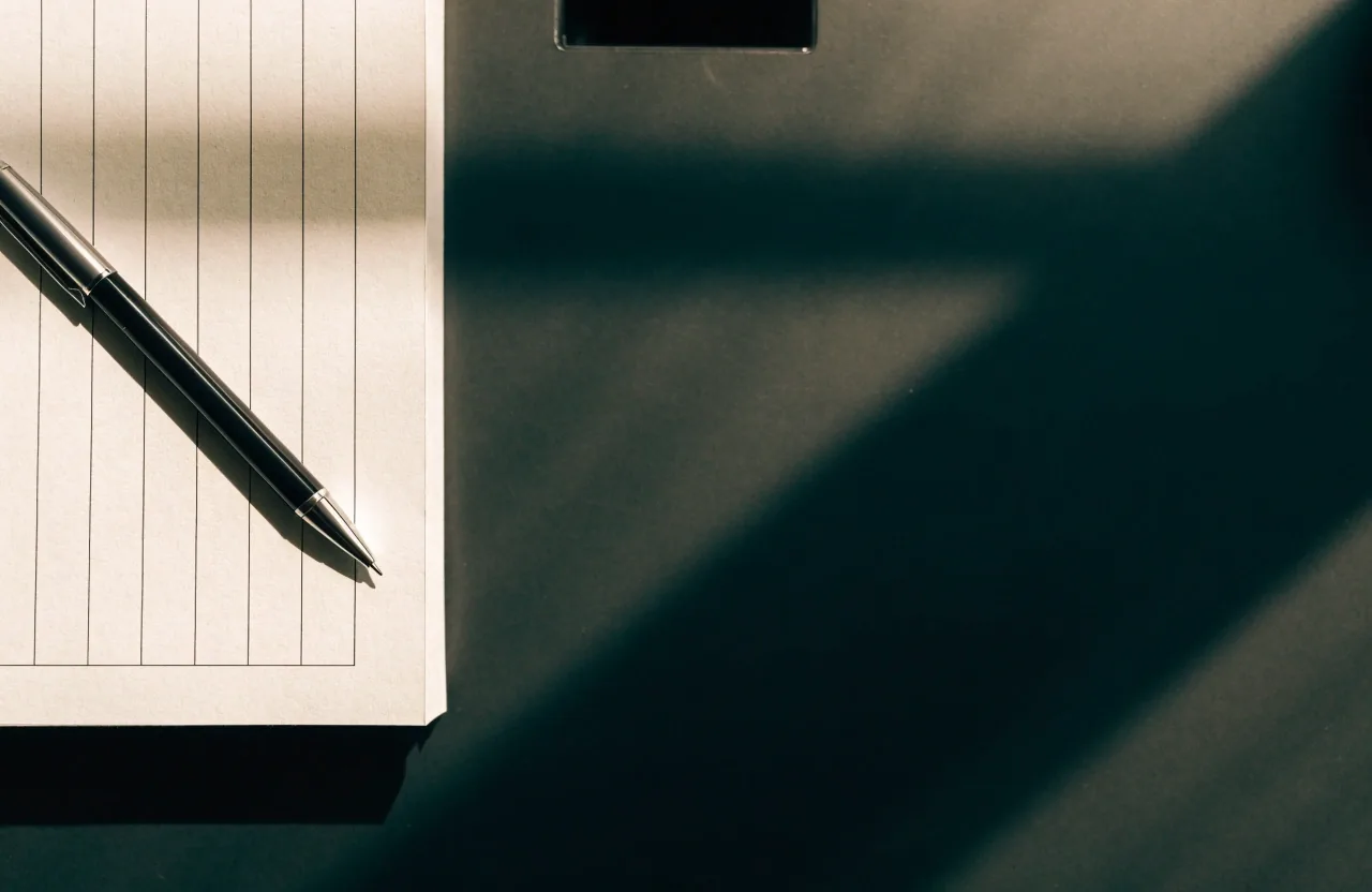 A pen resting across the ruled columns of an open ledger page on a dark desk under north skylight